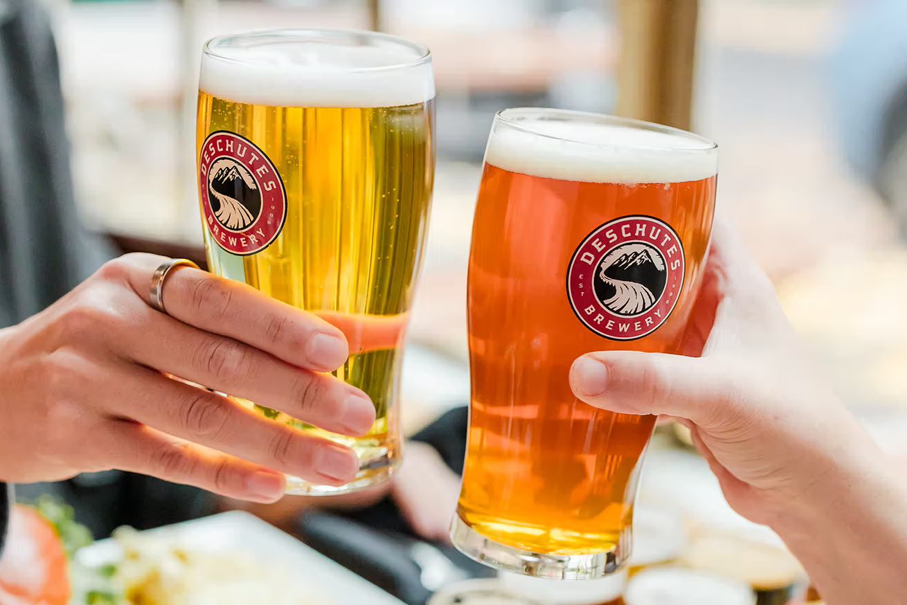 Two people clinking Deschutes Brewery beers in a bright restaurant scene, representing the Deschutes Brewery Try One on Us Instant Win Promotion where you can play daily to win an $8 PayPal or Venmo prize.