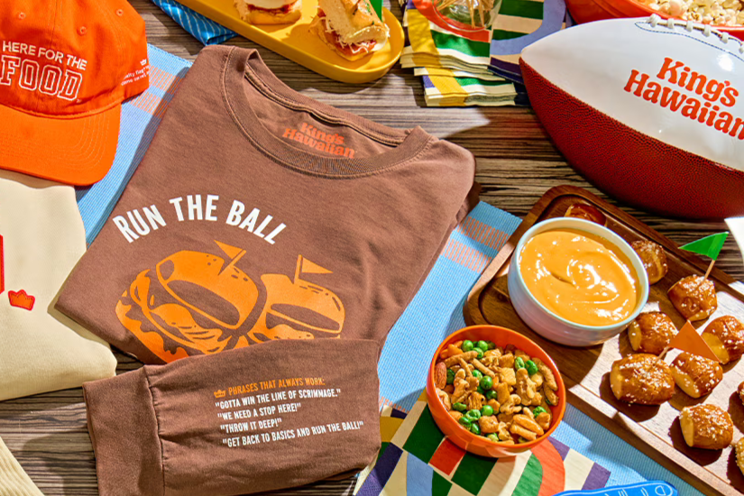 King’s Hawaiian football-themed prize image with a branded “Run the Ball” t-shirt, orange game-day hat, and a King’s Hawaiian football on a table, surrounded by party food like sliders, pretzels, dip, and snack mix; a King’s Hawaiian fan cave and football watch party giveaway prize spread featuring branded apparel and tailgate-style snacks; football season sweepstakes prize photo showing King’s Hawaiian gear, game-day food, and fan essentials.