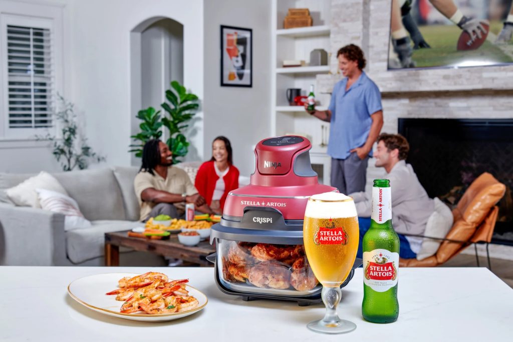 Lifestyle photo of a Stella Artois Hosting Worth More giveaway prize on a kitchen counter featuring a Stella Artois x Ninja Crispi air fryer with food inside, a Stella Artois beer bottle, and a branded beer glass, showing the Ninja Crispi prize package as the main sweepstakes giveaway item.