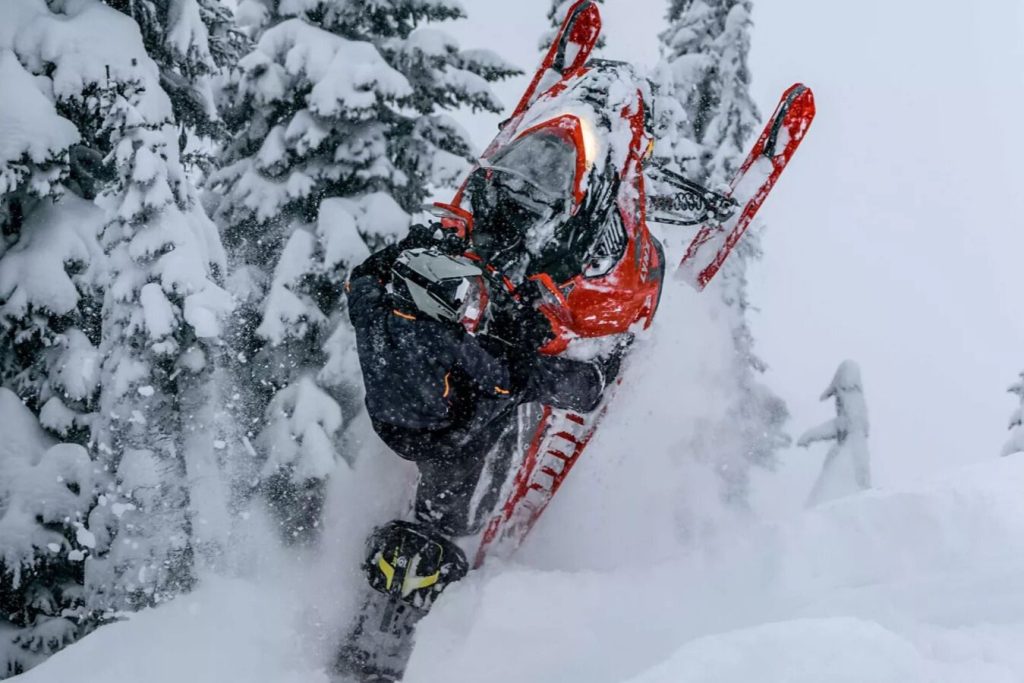 Action photo of a rider popping a bright red Lynx snowmobile up through deep powder in a snowy evergreen forest, capturing a high-energy Lynx snowmobile moment for the Win a Lynx snowmobile sweepstakes; dramatic winter riding scene highlighting a Lynx sled, snow spray, and backcountry conditions for a Lynx giveaway prize image.