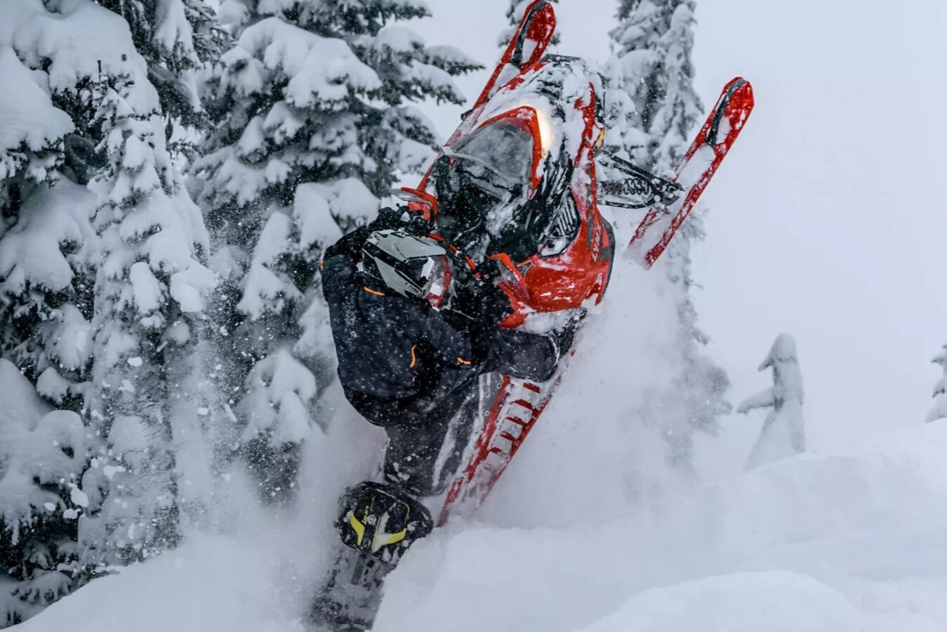 Action photo of a rider popping a bright red Lynx snowmobile up through deep powder in a snowy evergreen forest, capturing a high-energy Lynx snowmobile moment for the Win a Lynx snowmobile sweepstakes; dramatic winter riding scene highlighting a Lynx sled, snow spray, and backcountry conditions for a Lynx giveaway prize image.