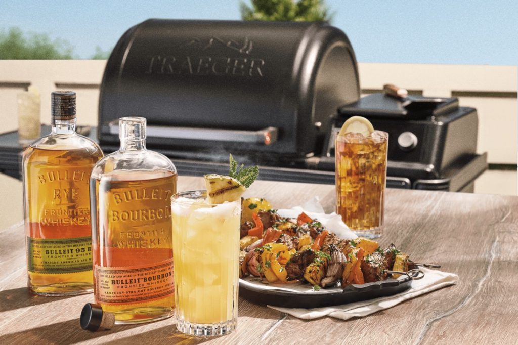 Bulleit Make The Meal Sweepstakes prize image featuring Bulleit Bourbon and Bulleit Rye bottles beside a cocktail, grilled skewers, and a Traeger grill, showing a Traeger Ironwood wood pellet grill and smoker prize bundle with a $250 e-gift card giveaway.