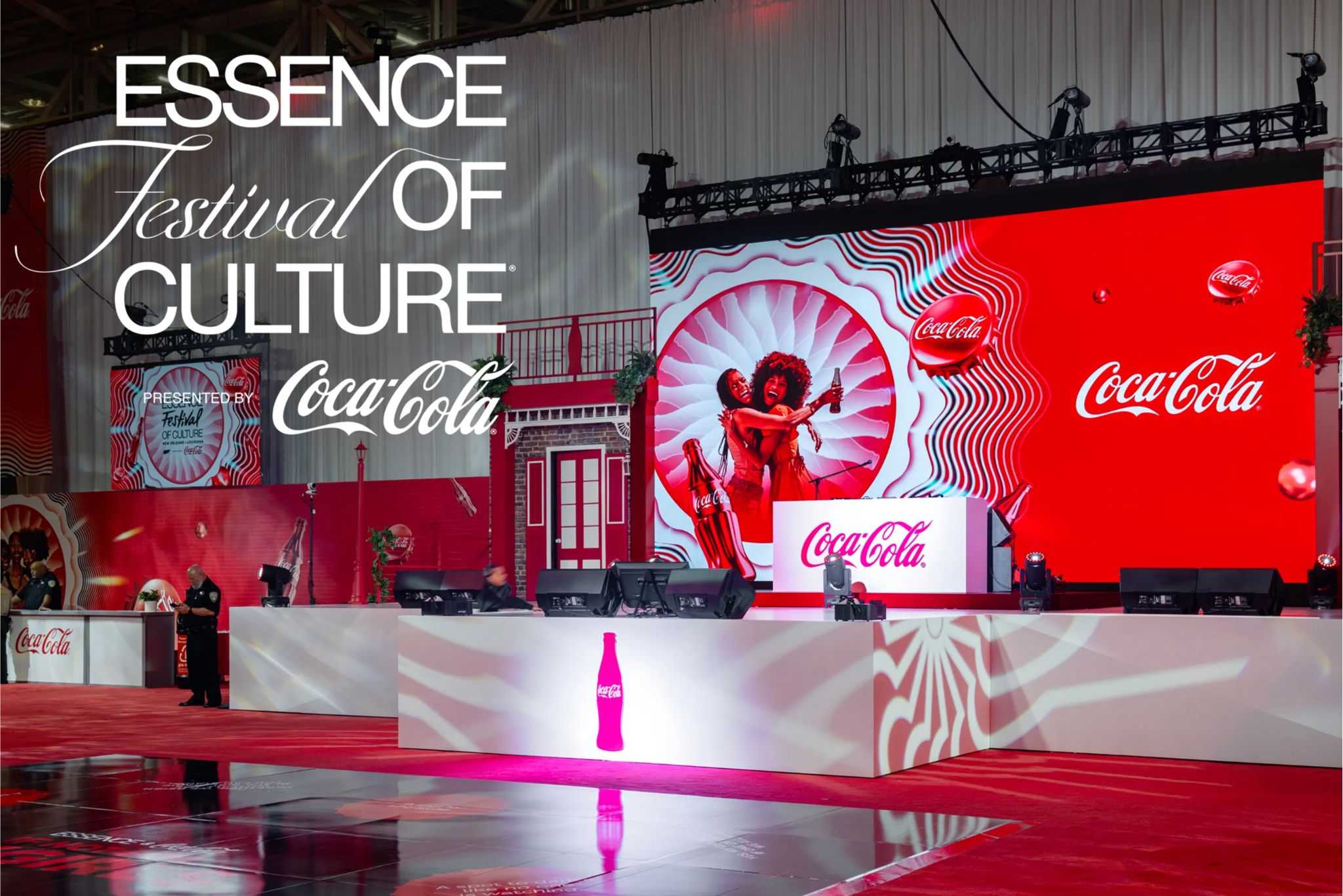 A large Coca-Cola branded stage display at the ESSENCE Festival of Culture featuring a bright red backdrop, Coca-Cola logos, and a promotional setup highlighting a New Orleans festival trip prize. The image shows a vibrant event stage with Coca-Cola branding tied to the ESSENCE Festival, representing a sweepstakes trip to New Orleans including festival passes and travel. The scene captures the energy of a major cultural music festival prize experience sponsored by Coca-Cola.