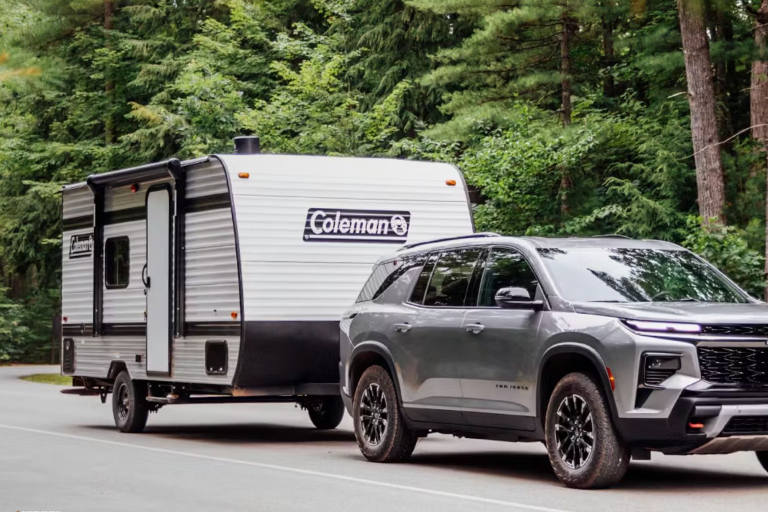A silver SUV towing a 2026 Keystone Coleman travel trailer on a forest road, showing the Camping World Win a New RV Giveaway prize. Coleman RV sweepstakes image featuring a modern tow vehicle and a white Coleman trailer, a new RV giveaway with a Keystone Coleman 13B travel trailer as the grand prize. Outdoor camping sweepstakes photo highlighting a Coleman camper trailer prize, RV giveaway promotion, and road-trip camping trailer sweepstakes theme.