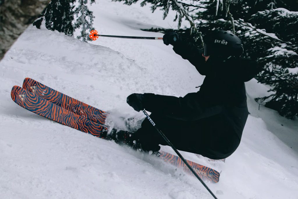 Action photo of a skier carving through deep powder in the trees on bright orange-and-blue patterned skis, representing the Fischer Ranger 102 LTD ski prize pack sweepstakes; winter ski giveaway featuring Ranger 102 LTD skis plus Ford Ranger swag like YETI cooler gear, duffel, blanket, and trucker hat.