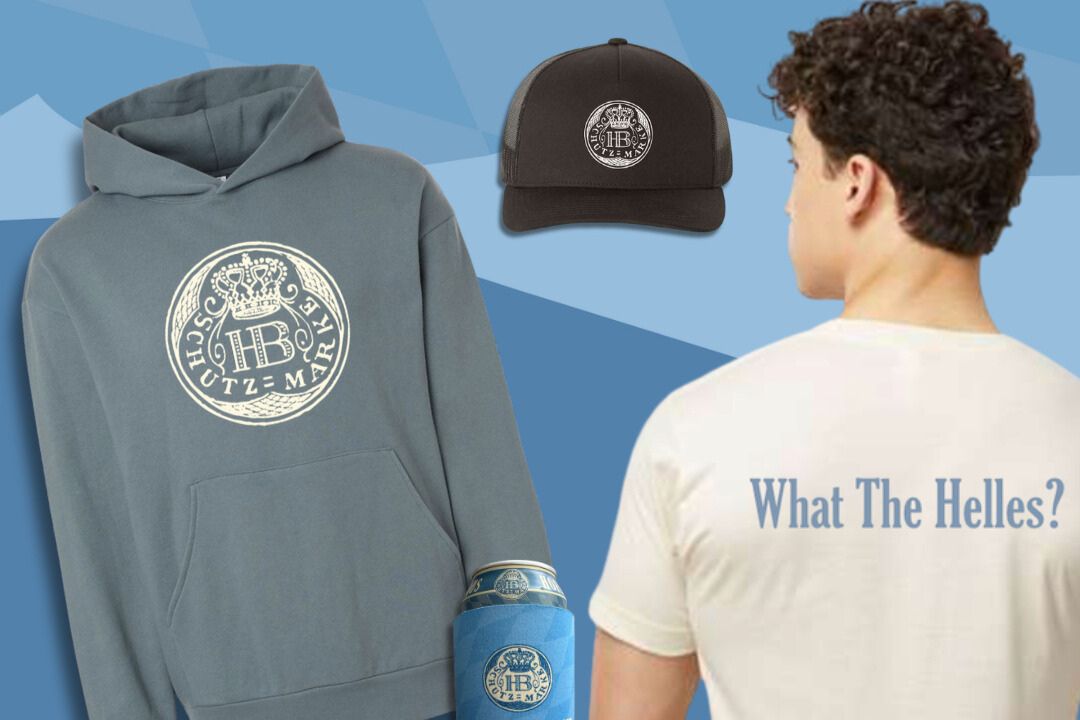 A Hofbräuhaus Helles swag prize bundle featuring a branded hoodie, a black trucker hat, a blue koozie, and a “What the Helles” t-shirt shown on a model, all included in the Hofbräuhaus Helles Swag National Sweepstakes. The image highlights the giveaway prize pack with Hofbräuhaus Helles Schutzmarke logo apparel and accessories, showing the hoodie graphic, the hat logo, and the shirt back print in one clear sweepstakes prize display. This Hofbräuhaus sweepstakes prize photo focuses on the merch kit you can win, including the hoodie, t-shirt, hat, and koozie, as part of the Helles gear swag giveaway.