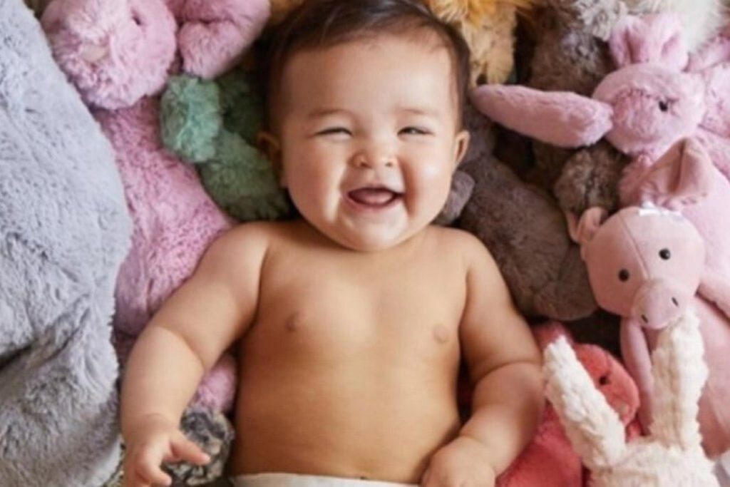 Smiling baby lying on a blanket surrounded by plush stuffed animals and soft toys, cozy nursery-style scene that fits a Huggies diapers for a year sweepstakes prize, cute infant photo representing baby essentials, diapers, parenting giveaways, and a year supply of Huggies diaper prize.