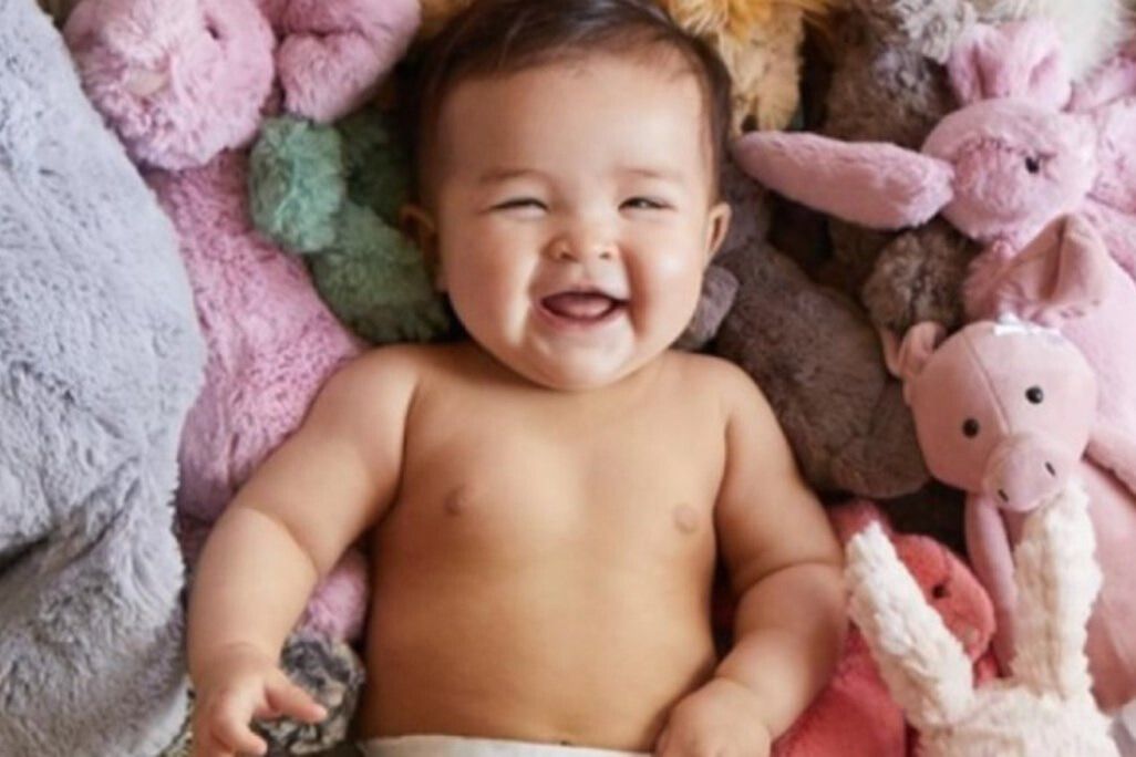 Smiling baby lying on a blanket surrounded by plush stuffed animals and soft toys, cozy nursery-style scene that fits a Huggies diapers for a year sweepstakes prize, cute infant photo representing baby essentials, diapers, parenting giveaways, and a year supply of Huggies diaper prize.