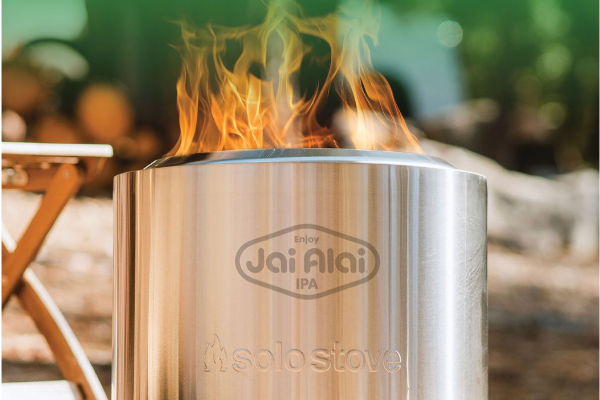 Close-up promotional photo of a stainless steel Solo Stove Bonfire fire pit with flames rising from the top and a Jai Alai IPA logo etched on the side, showing an outdoor fire pit prize for the Jai Alai Hat Trick Sweepstakes and highlighting a Solo Stove giveaway for backyard and camping-style fires.
