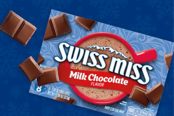 Close-up image of a Swiss Miss Milk Chocolate hot cocoa box on a blue background with chocolate squares around it, showing the Swiss Miss hot cocoa mix packaging; Swiss Miss sweepstakes image featuring the Milk Chocolate flavor box and chocolate pieces, a cozy cocoa-themed giveaway visual for a Swiss Miss review and win promotion.