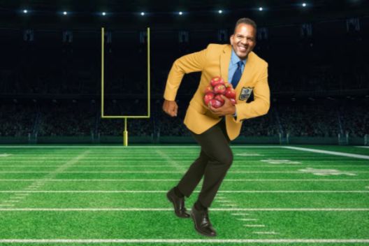 Man in a gold football-style jacket running on a football field holding a bundle of red apples, promoting the SnapDragon Apples sweepstakes. Snap Your Way to Canton Giveaway image suggesting a Pro Football Hall of Fame trip prize with SnapDragon apples branding. Sweepstakes graphic for a chance to win a Canton, Ohio football experience trip, featuring SnapDragon apples and a stadium field background.