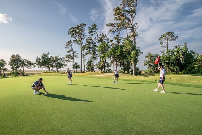 Four adults stand on a manicured coastal golf green in the Golden Isles lining up a putt near the flagstick, with oceanfront trees and blue sky in the background. The image shows a scenic Georgia golf course tied to a Golden Isles getaway giveaway featuring a beach resort stay and golf experience.