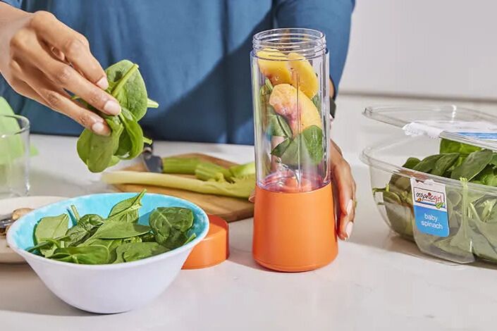 A person adding fresh baby spinach to a blender cup filled with fruit on a kitchen counter, prepping a green smoothie with leafy greens and produce. Fresh spinach and smoothie ingredients in a personal blender, a healthy greens kitchen scene that fits an organicgirl giveaway vibe. Close-up of spinach, fruit, and a small blender cup—fresh salad-style greens and smoothie prep imagery tied to the organicgirl sweepstakes featuring an Expedia gift card and a year of free organicgirl salads.