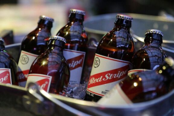 Six brown Red Stripe beer bottles sitting in a metal ice bucket at a bar. Chilled Red Stripe lager bottles in ice, a close-up of Red Stripe beer labels, a beer for a year sweepstakes prize-style photo.