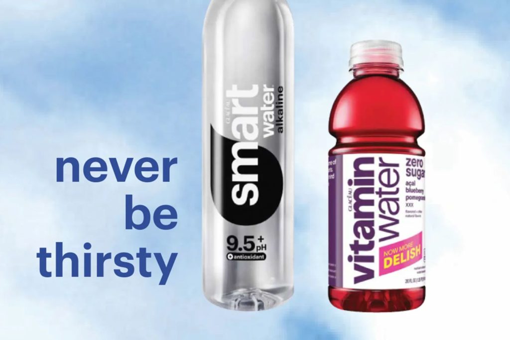 A smartwater bottle next to a vitaminwater zero sugar bottle on a blue background with the words “never be thirsty,” representing the smartwater vitaminwater Product for a Year Sweepstakes grand prize. The image highlights a year of hydration products with monthly deliveries of smartwater or vitaminwater. A smartwater and vitaminwater giveaway visual showing the sweepstakes prize of 24 bottles per month for 12 months.