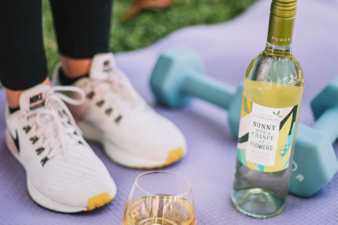 A close-up photo of a purple yoga mat with teal dumbbells, a pair of white running shoes, and a bottle of Sunny with a Chance of Flowers wine beside a glass; the shot shows workout gear and a wine bottle staged on a mat to suggest fitness and lifestyle. A fitness-themed image featuring sneakers, a yoga mat, light dumbbells and a Sunny with a Chance of Flowers wine bottle, emphasizing a workout-and-reward vibe and connecting the brand to active living. Promotional imagery pairing exercise props and Sunny wine that highlights the Resolution Workout Wardrobe Sweepstakes prize theme of yoga and fitness accessories.