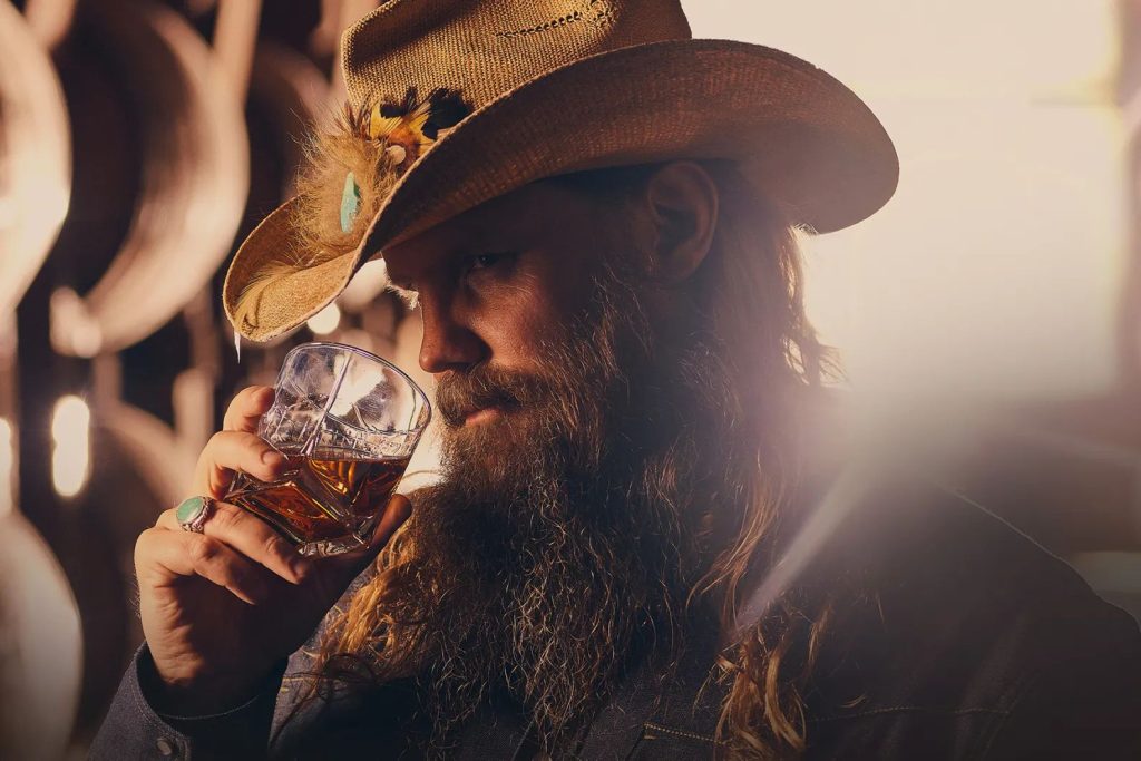 A bearded man in a cowboy hat holds a glass of whiskey in a warm-lit setting, representing the Traveller Whiskey Made for the Moment Sweepstakes grand prize concert and VIP travel experiences. The image shows a rugged country-style atmosphere with a whiskey glass in hand, symbolizing the music-inspired prize packages tied to Chris Stapleton concerts and exclusive event trips. A close-up of a man enjoying whiskey captures the vibe of the Traveller Whiskey Sweepstakes featuring concert tickets, signed memorabilia, and VIP travel experiences.