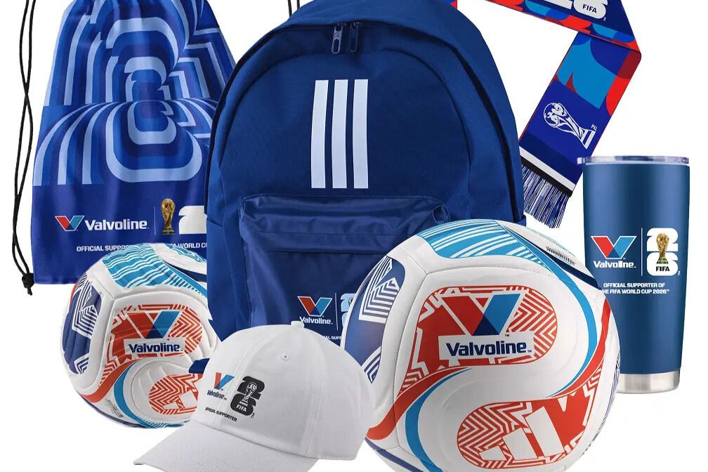 A promotional image showing Valvoline-branded FIFA merchandise including two full-size soccer balls, a mini soccer ball, a blue backpack, a drawcord bag, a baseball cap, a scarf, and a 20oz tumbler, representing the Instant Win prizes in the Valvoline FIFA World Cup Sweepstakes. The image highlights soccer-themed prize items tied to the Valvoline For the Driven Promotion, featuring official FIFA World Cup merchandise such as balls, bags, and drinkware. A collection of Valvoline FIFA giveaway prizes displayed together, including sports gear and branded accessories connected to the 2026 FIFA World Cup Final trip sweepstakes.