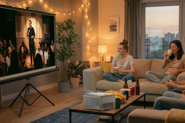 Cozy living room movie night with friends watching a stand-up comedy show on a projector screen, matching a BenQ projector giveaway prize for at-home entertainment. Warm string lights, snacks, and a projector setup highlight a home theater vibe that fits the BenQ Ready for Better Moments Together campaign. Home entertainment projector giveaway image showing a projector screen in a modern apartment, emphasizing a BenQ projector prize bundle for movie nights.
