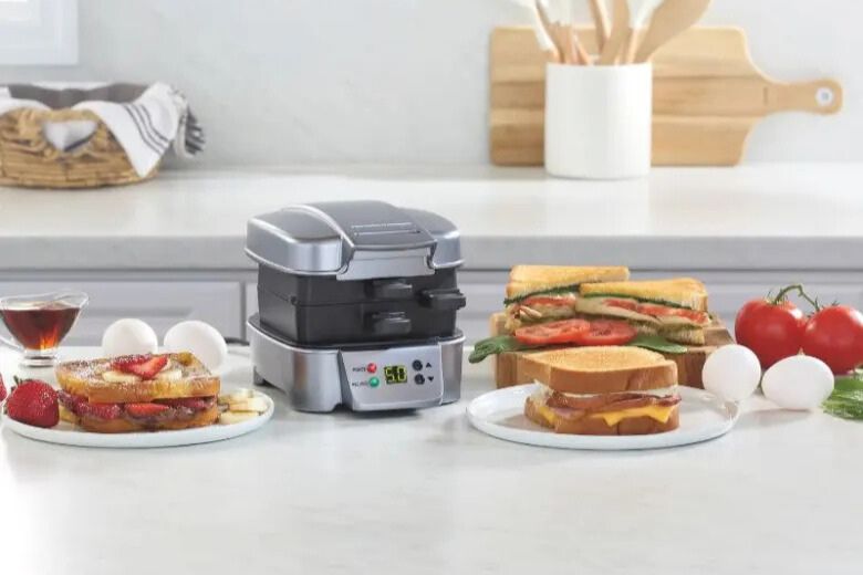 Kitchen countertop photo showing a Hamilton Beach Sandwich Stacker hot sandwich maker in silver with prepared breakfast sandwiches, eggs, tomatoes, and toast nearby, highlighting the giveaway prize appliance. The image features the compact hot sandwich maker used for stacked breakfast sandwiches and grilled sandwiches, showing a Hamilton Beach sandwich maker sweepstakes prize in a bright modern kitchen setting.