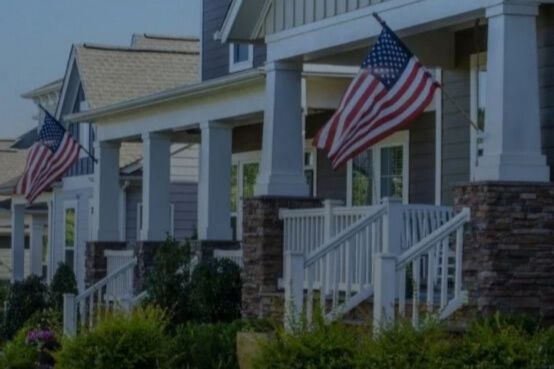 A row of suburban American homes with front porches and large U.S. flags hanging outside, illustrating the Realtor.com $75K Sweepstakes and Veterans United Home Loans 2026 New Year, New Home $75K Veteran Homebuyer Giveaway for a $75,000 home purchase prize.