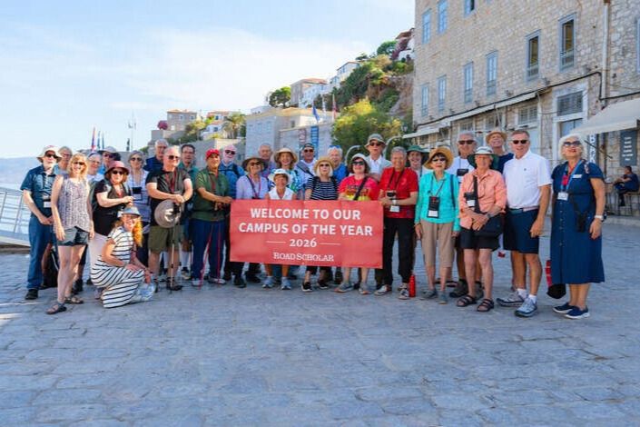 A large group of adult travelers stands together on a sunny waterfront in Greece holding a banner that reads Welcome to Our Campus of the Year 2026 Road Scholar. The photo shows participants on an educational travel tour in a historic Greek coastal town, representing the type of learning adventure featured in the Road Scholar Greece Trip Sweepstakes prize. Travelers on a guided cultural tour gather near stone buildings and a harbor, illustrating the immersive travel experience offered in the Greece educational travel sweepstakes.