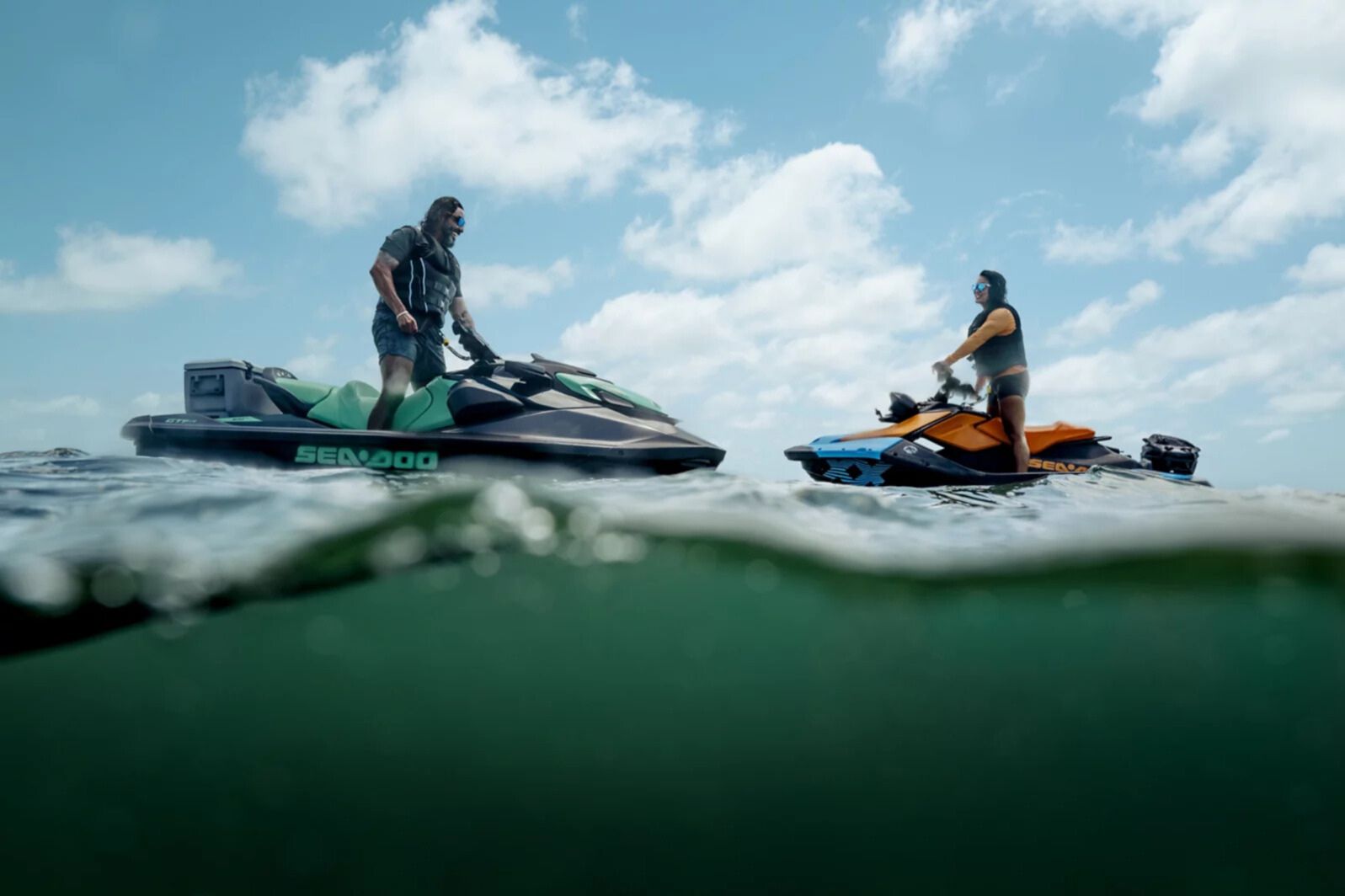 Split-level ocean photo showing two riders on Sea-Doo personal watercraft facing each other on the water under a bright blue sky with clouds. The image highlights a Sea-Doo sweepstakes prize theme with personal watercraft on open water. It visually represents a BRP vehicle giveaway featuring Sea-Doo jet skis and adventure-style riding. v