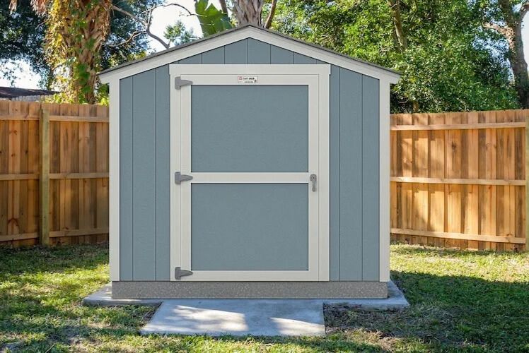 A gray and white Tuff Shed-style backyard storage shed with double doors sitting on a concrete pad inside a fenced yard with trees behind it. Backyard storage shed prize image showing an 8x8 style outdoor shed structure, clean modern panels, and a simple garden setting. Outdoor shed giveaway photo featuring a compact storage building in a residential backyard, representing a shed sweepstakes prize.