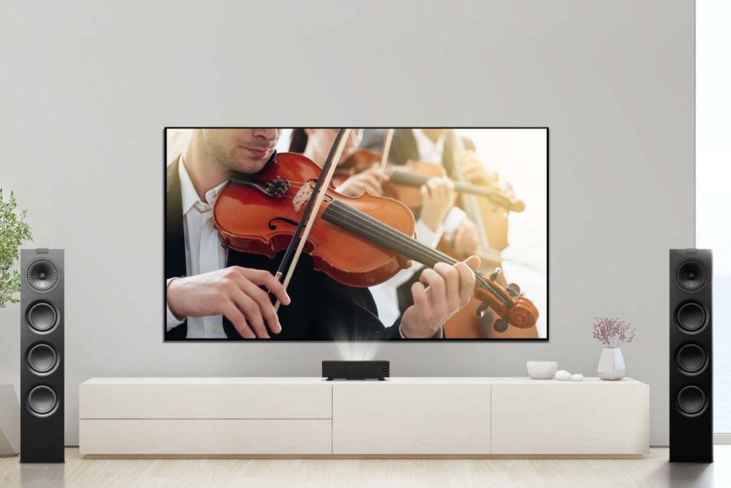 A living room features a large screen displaying a musician playing a violin, while other musicians are blurred in the background. Stylish speakers flank a modern entertainment unit, featured in the BenQ Director's Cut Sweepstakes