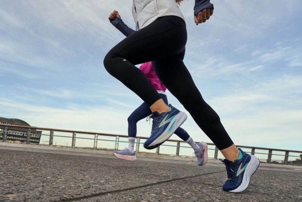 Two runners in athletic wear are sprinting along a paved path, with one wearing navy shoes and the other in pastel colors, against a backdrop of blue skies and distant buildings, featured in the Brooks Running Race Giveaway