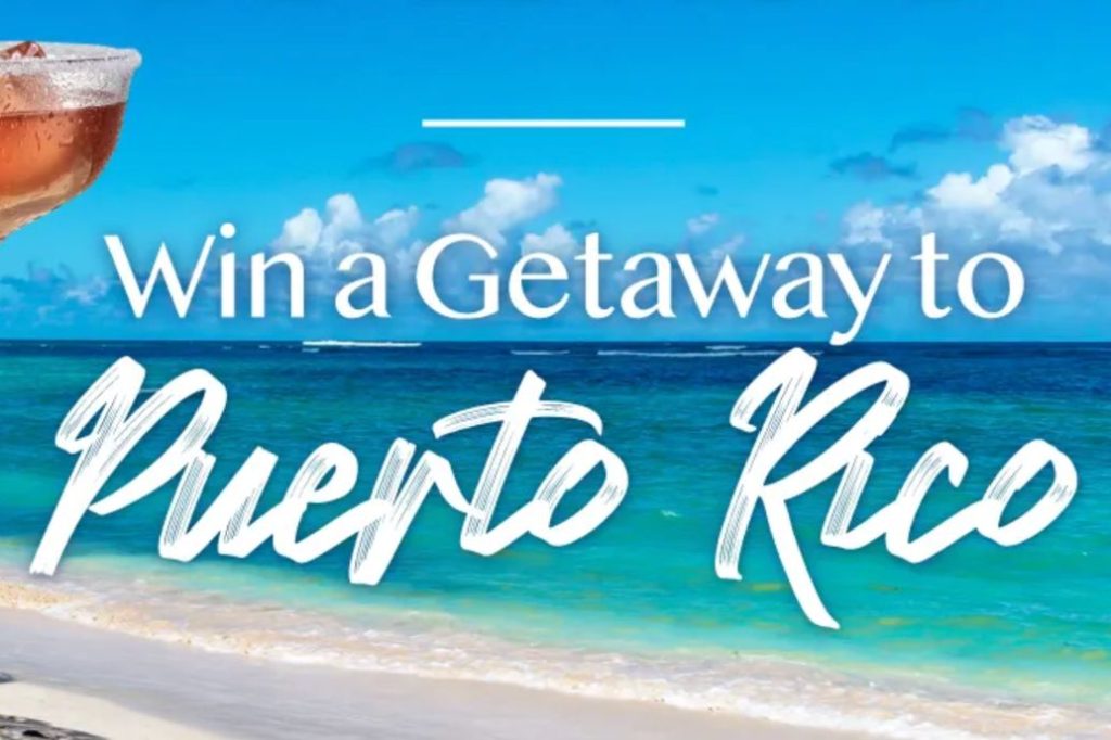 A tropical beach scene features a clear blue sky, turquoise waters, and soft sand stretching along the shore, creating a serene vacation atmosphere, featured in the PAMA in Paradise Sweepstakes