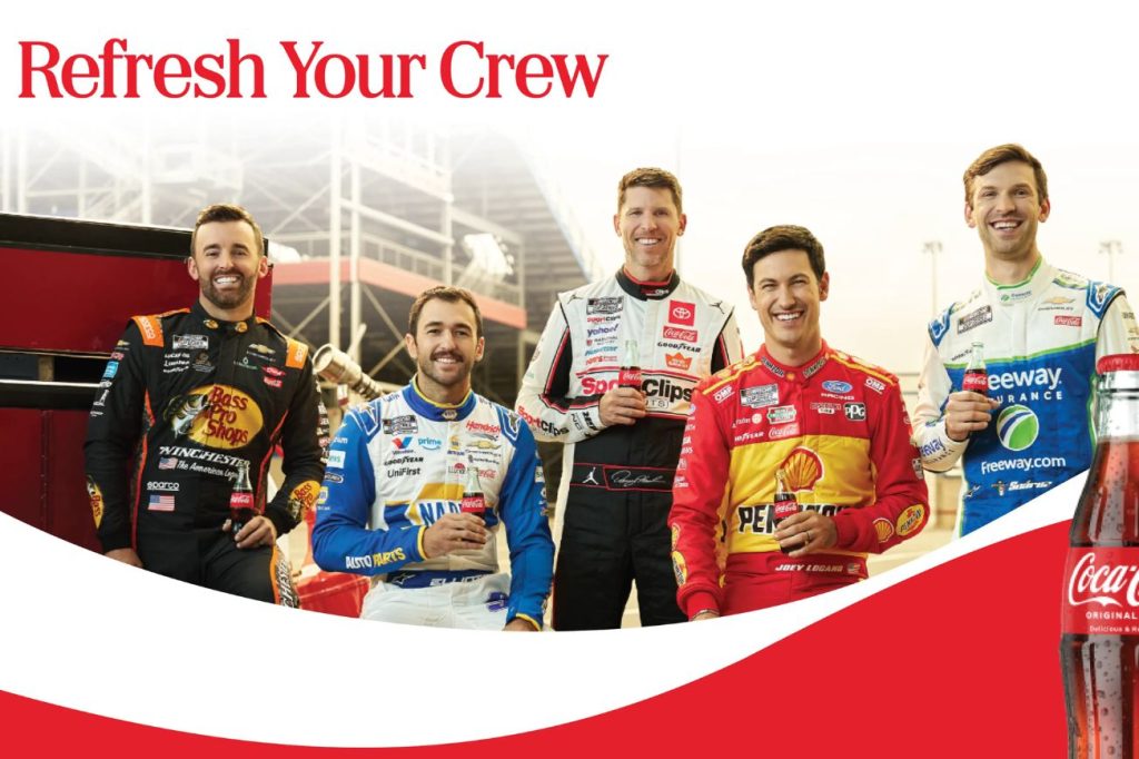 Five smiling race car drivers in team uniforms stand together, each holding a beverage, against a racetrack backdrop, showcasing camaraderie and celebration, featured in the Coca Cola Flyaway Sweepstakes