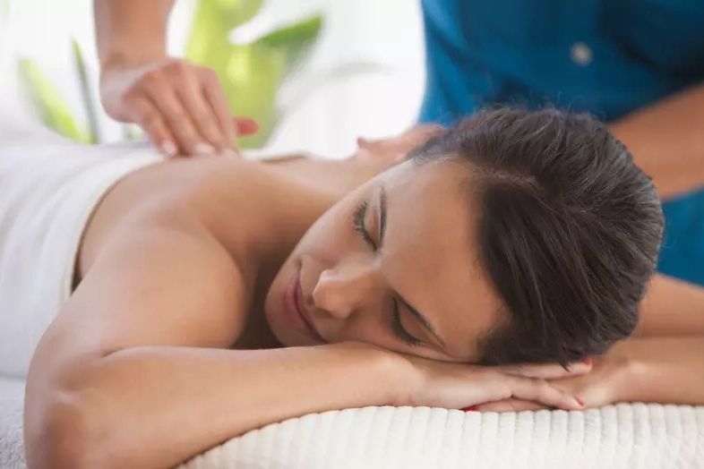 A woman relaxes on a massage table while receiving a back massage. The serene setting features soft lighting and green plants in the background, featured in the Massage Envy Scholarship Sweepstakes