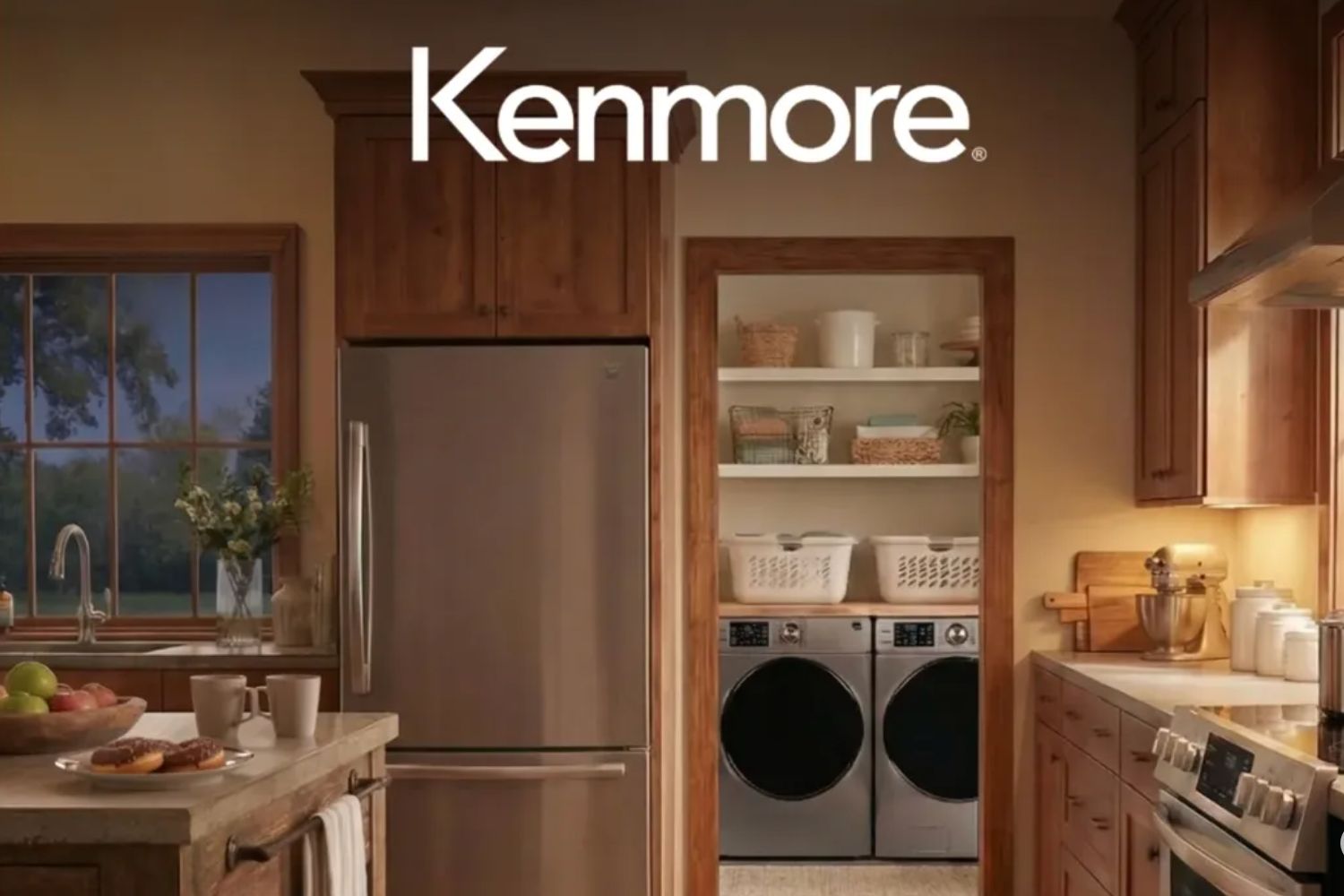 A cozy kitchen features a stainless steel refrigerator, a wooden island with fresh fruit and pastries, and a laundry area with stacked machines visible in the background, featured in the Kenmore Appliance Refresh Sweepstakes