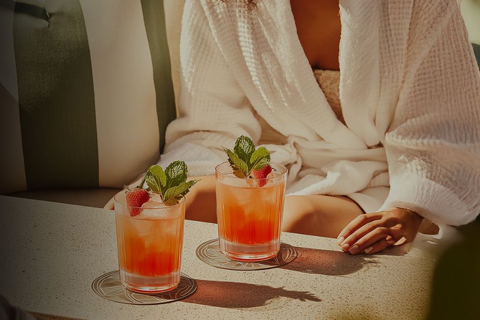 A woman in a white bathrobe relaxes by a poolside, gently resting her hand on the table, which features two vibrant cocktails garnished with mint and strawberries, featured in the Ghost Tequila Spa Day Sweepstakes