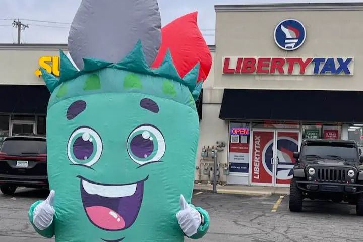 A cheerful inflatable Statue of Liberty mascot stands in front of a tax service office, arms outstretched and smiling, with parked cars visible in the background, featured in the Liberty Tax $300 Sweepstakes