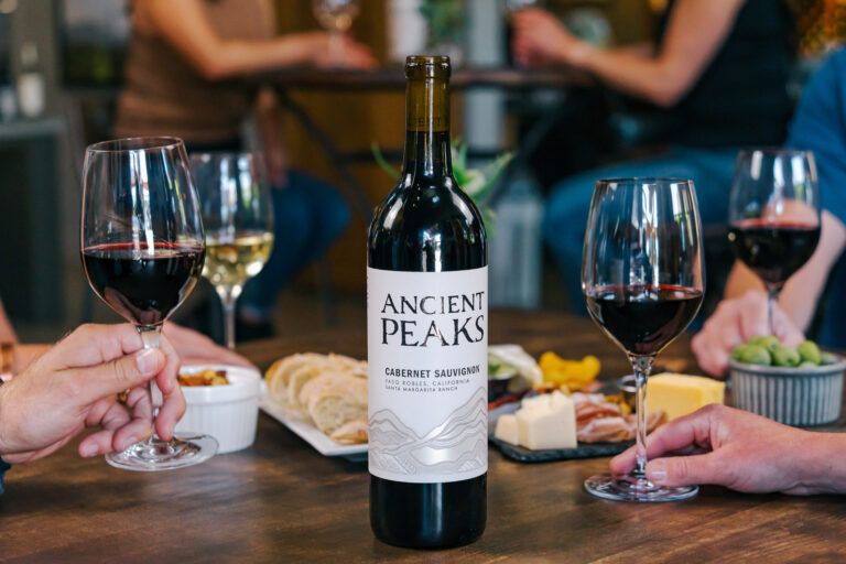 A bottle of Cabernet Sauvignon sits on a table surrounded by glasses of red and white wine, with friends enjoying snacks like cheese and olives in a warm, social setting, featured in the Ancient Peaks Trip Sweepstakes