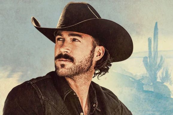 A man with a mustache and long hair wears a wide-brimmed cowboy hat and a dark jacket, gazing thoughtfully at the horizon against a muted desert background, featured in the Black Rifle Riley Green VIP Sweepstakes