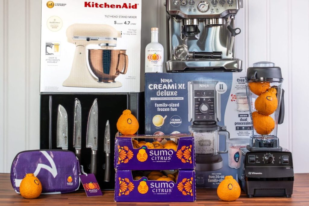 A collection of kitchen appliances and tools, including a stand mixer, espresso machine, and blender, alongside two crates of Sumo citrus oranges and kitchen knives, featured in the Sumo Citrus Taste of Joy Sweepstakes
