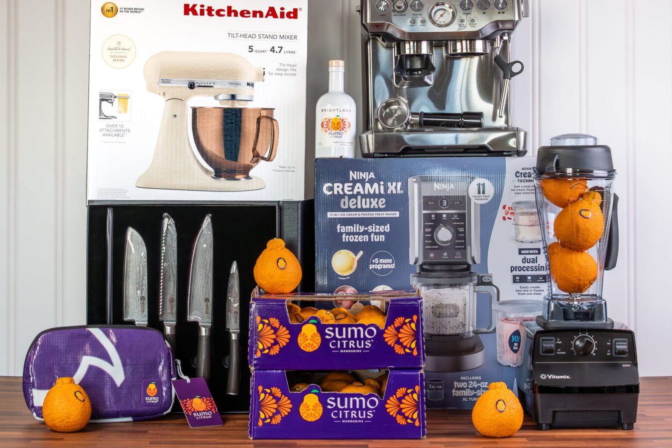 A collection of kitchen appliances and tools, including a stand mixer, espresso machine, and blender, alongside two crates of Sumo citrus oranges and kitchen knives, featured in the Sumo Citrus Taste of Joy Sweepstakes