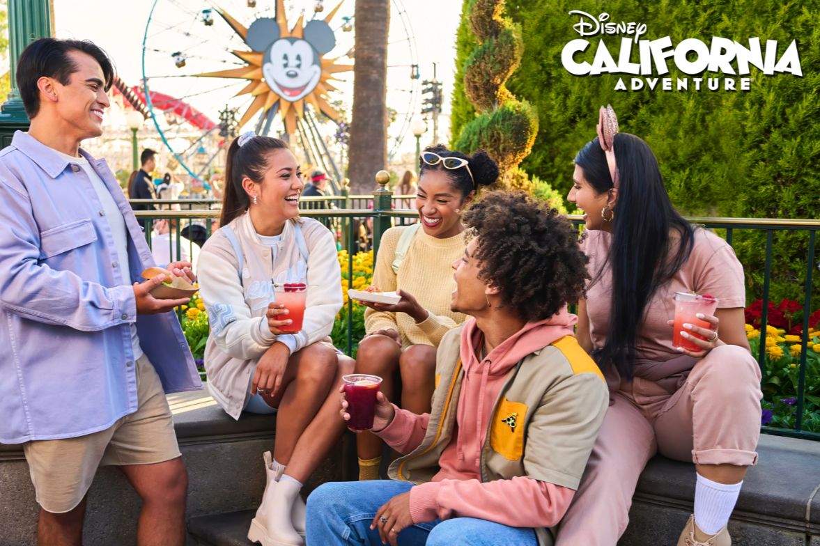 A group of five friends enjoys snacks and colorful drinks while laughing together near vibrant flowers and a large Mickey Mouse-themed attraction in the background, featured in the The Woody Show After Hours Takeover Sweepstakes