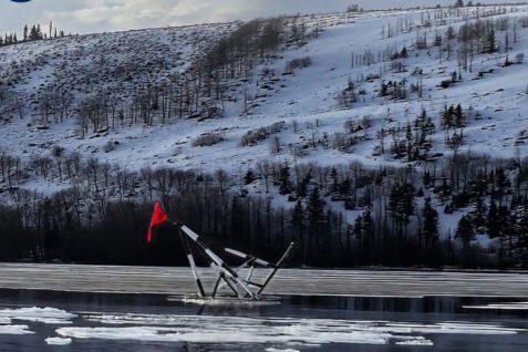 A metal structure with a red flag is partially submerged in a frozen lake, surrounded by snow-covered hills and sparse trees under a cloudy sky, featured in the Holland America 2026 Alaska Sweepstakes