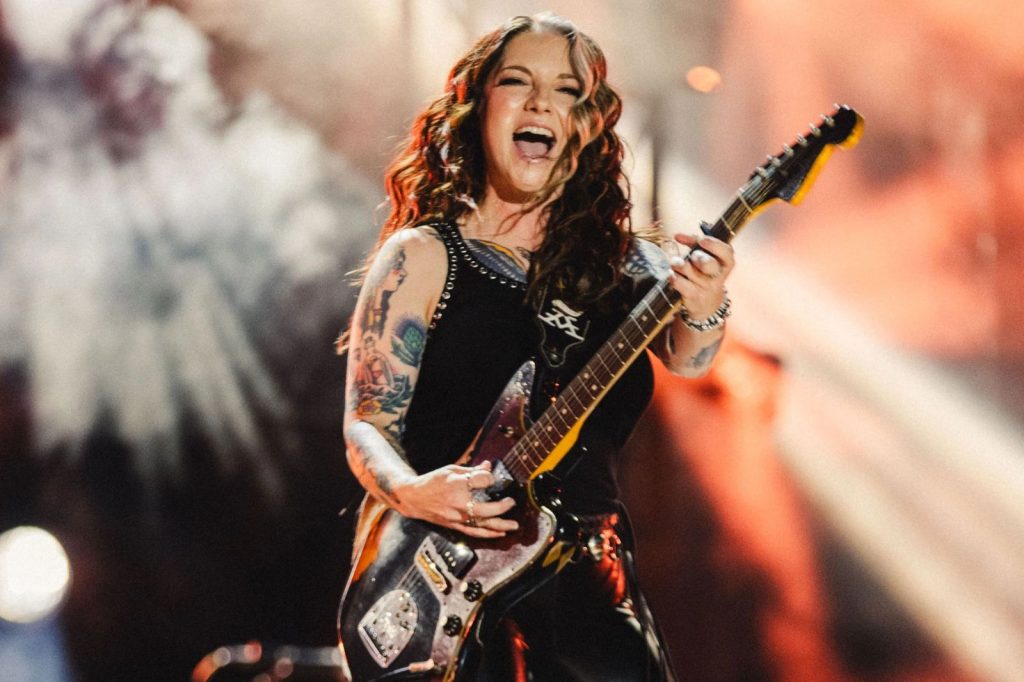 A musician passionately plays an electric guitar on stage, smiling widely with long, wavy hair, wearing a sleeveless top and leather pants, while vibrant lights illuminate the background, featured in the Ashley McBryde Live in Music City Giveaway