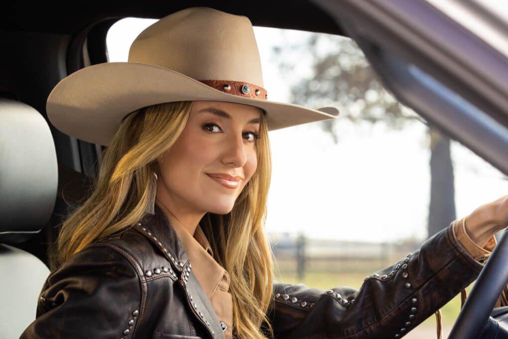 A woman with long blonde hair and a large beige cowboy hat smiles confidently while sitting in a vehicle, showcasing a relaxed countryside atmosphere, featured in the Ford Lainey Wilson Signed Guitar Sweepstakes