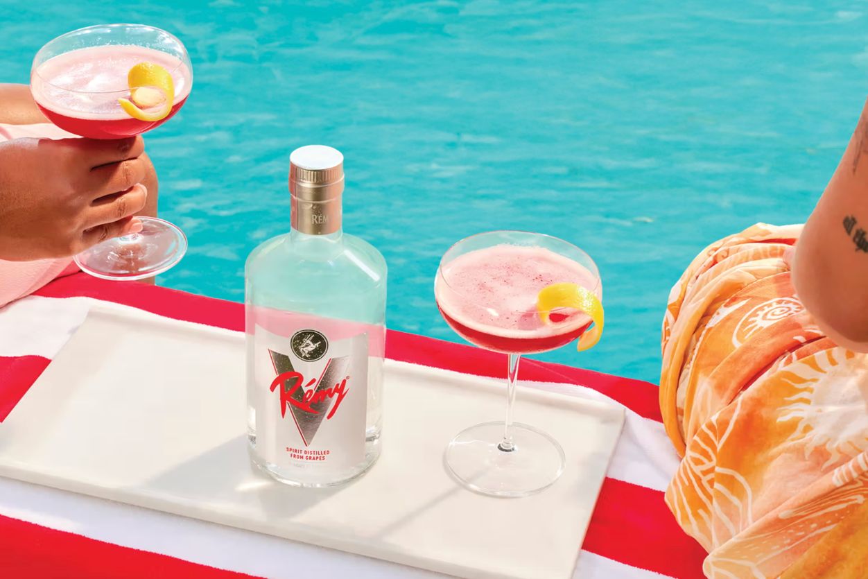 Bright cocktails with lemon garnishes rest on a tray by a shimmering pool, as hands enjoy drinks under clear blue skies, creating a vibrant summer atmosphere, featured in the Rémy V My Vibe Sweepstakes