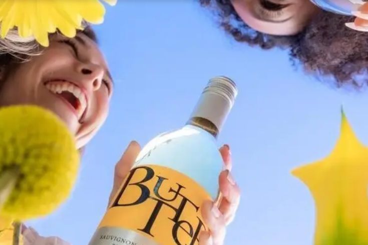 Two people enjoy a sunny day, holding a bottle of Sauvignon Blanc while surrounded by blooming flowers, smiling and showcasing the joyful atmosphere, featured in the JaM Cellars Year of Blooms Sweepstakes