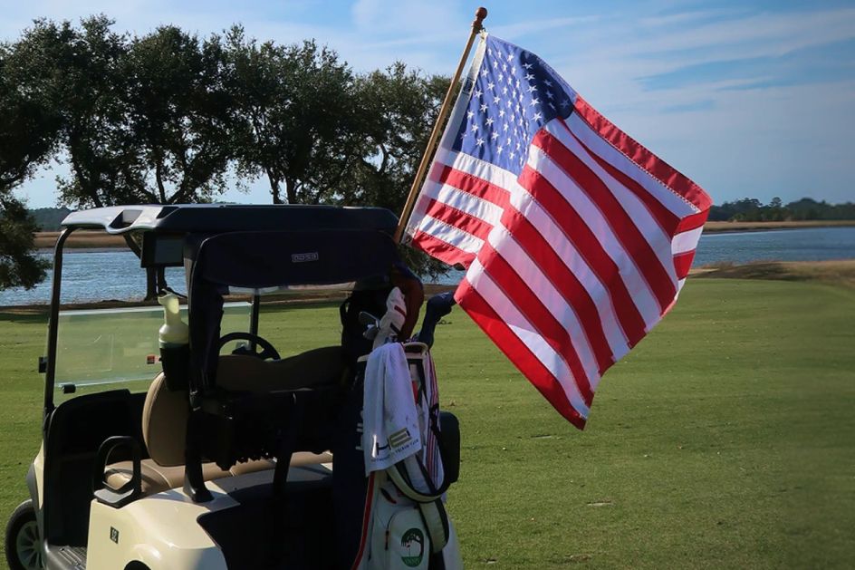 A golf cart is parked on the green, featuring a prominent American flag waving in the breeze. In the background, a serene waterway and trees create a picturesque setting, featured in the Allegiance PGA Championship Sweepstakes