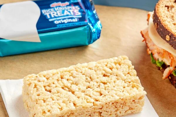 A crispy rice treat sits on a white napkin, next to a wrapped snack and a sandwich with layers of turkey, lettuce, and cheese, all on a brown paper surface, featured in the Kelloggs Solo Stove Sweepstakes