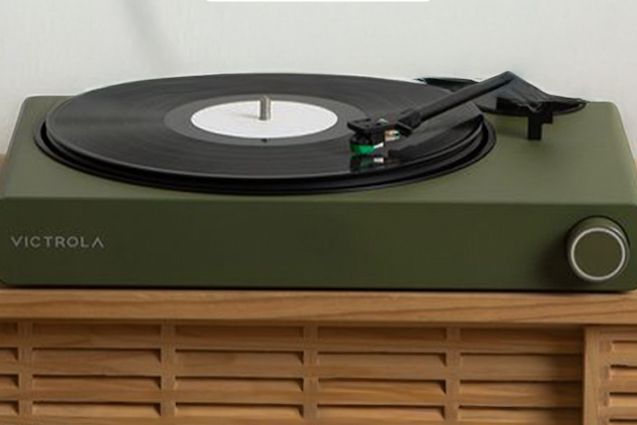 A vintage-style turntable in olive green rests on a wooden stand, with a black vinyl record spinning and the tonearm poised to play music, featured in the Victrola Stream Onyx & Vinyl Sweepstakes