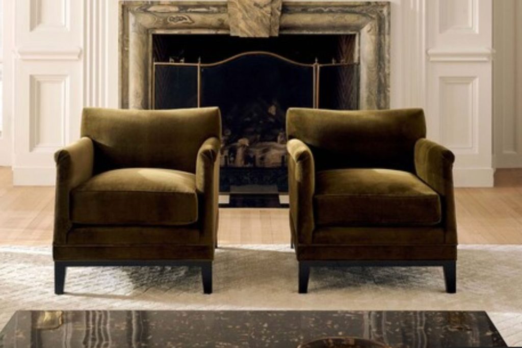 Two plush, olive-green armchairs are positioned symmetrically in a cozy room, facing a marble fireplace. Light wooden flooring complements the elegant, inviting space, featured in the CB2 $1,000 Gift Card Sweepstakes